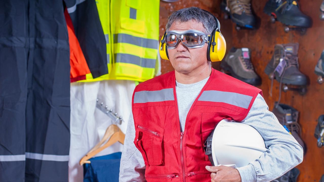 Industrial worker wearing hearing protection and a red vest, carrying a white helmet